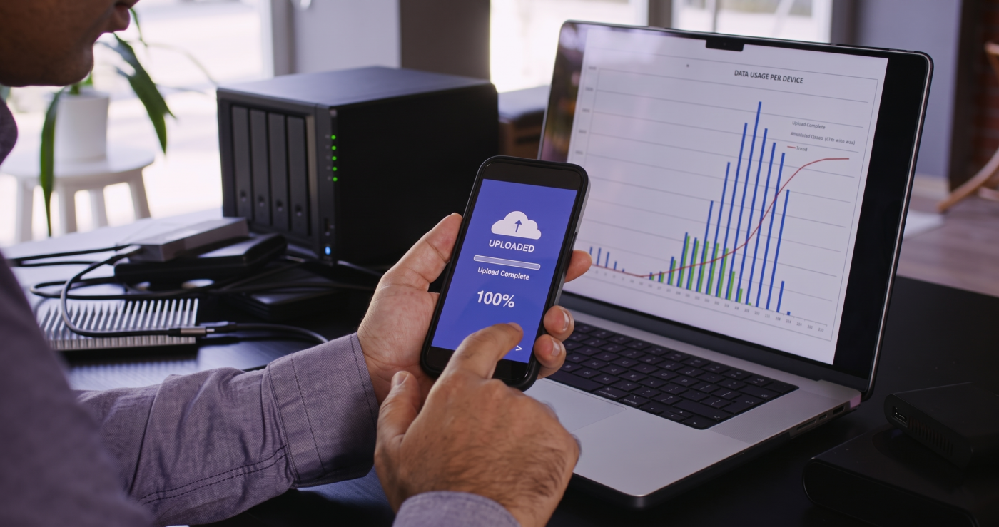 A business professional holding a smartphone displaying a 100 percent uploaded confirmation screen, sitting at a desk next to a laptop showing data usage graphs and a black network-attached storage backup drive.