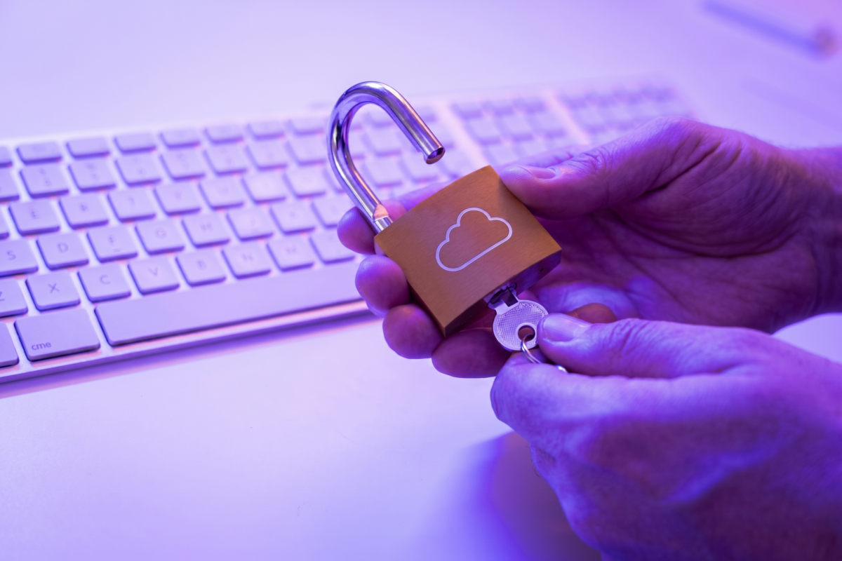 Two hands holding an unlocked brass padlock featuring a white cloud icon, hovering above a white computer keyboard under purple lighting, representing vulnerabilities in cloud security configurations.