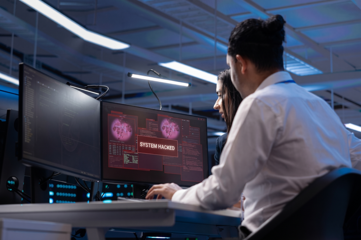 Two cybersecurity professionals in a dark operations center urgently looking at dual monitors displaying a red System Hacked warning alert during a network breach.